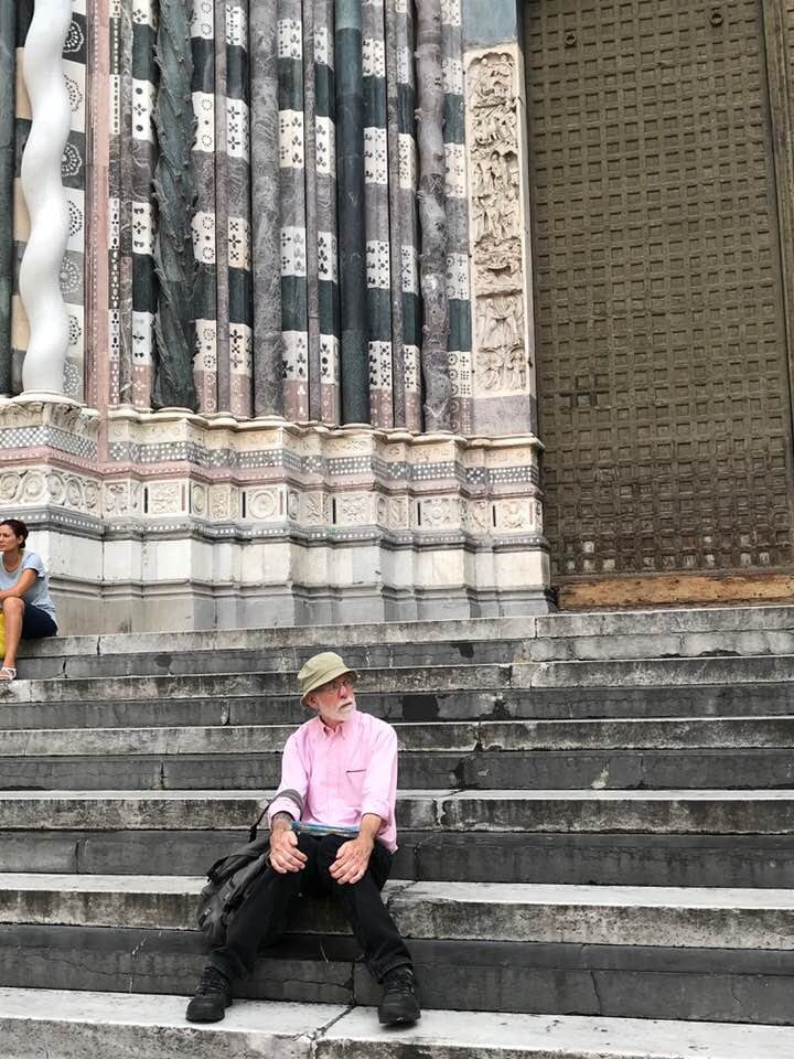 person with a gray beard, hat and glasses, holding a backpack sitting on the steps of the Duomo in Genoa, Italy
