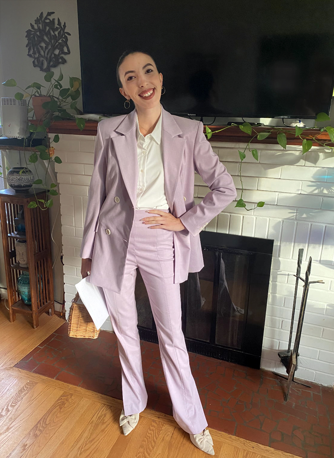 person with brown hair and earrings in a lavender suit standing in a living room, hand on hip and smiling